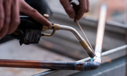 Hand holding a welding flux rod while heating it to solder a copper pipe and elbow